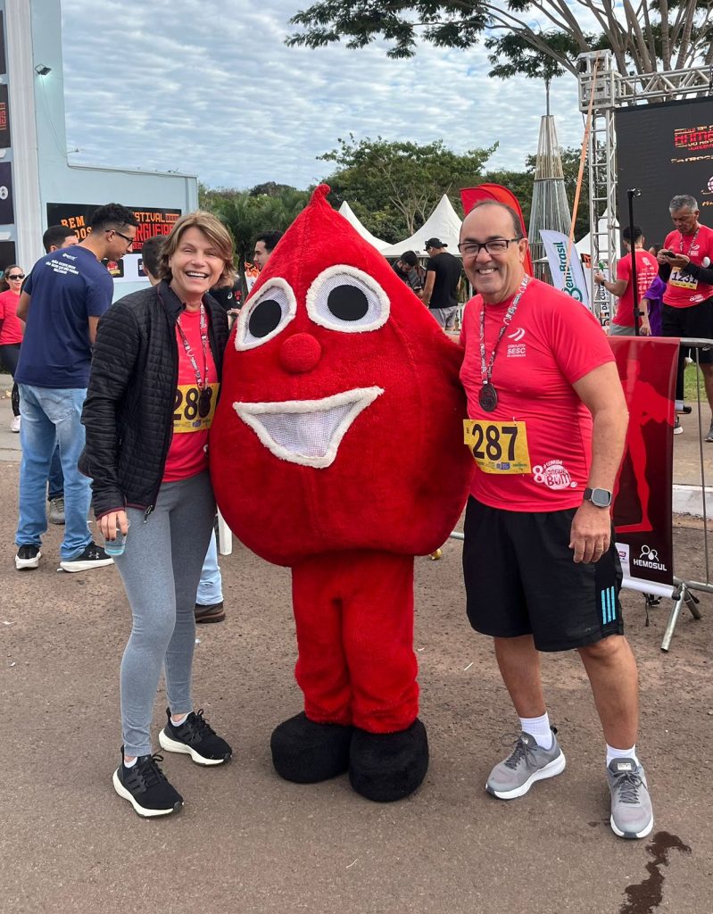 imagem com duas pessoas abrasando uma pessoa fantasiada com traje de gota de sangue, em referencia a consientização a doação de sangue.